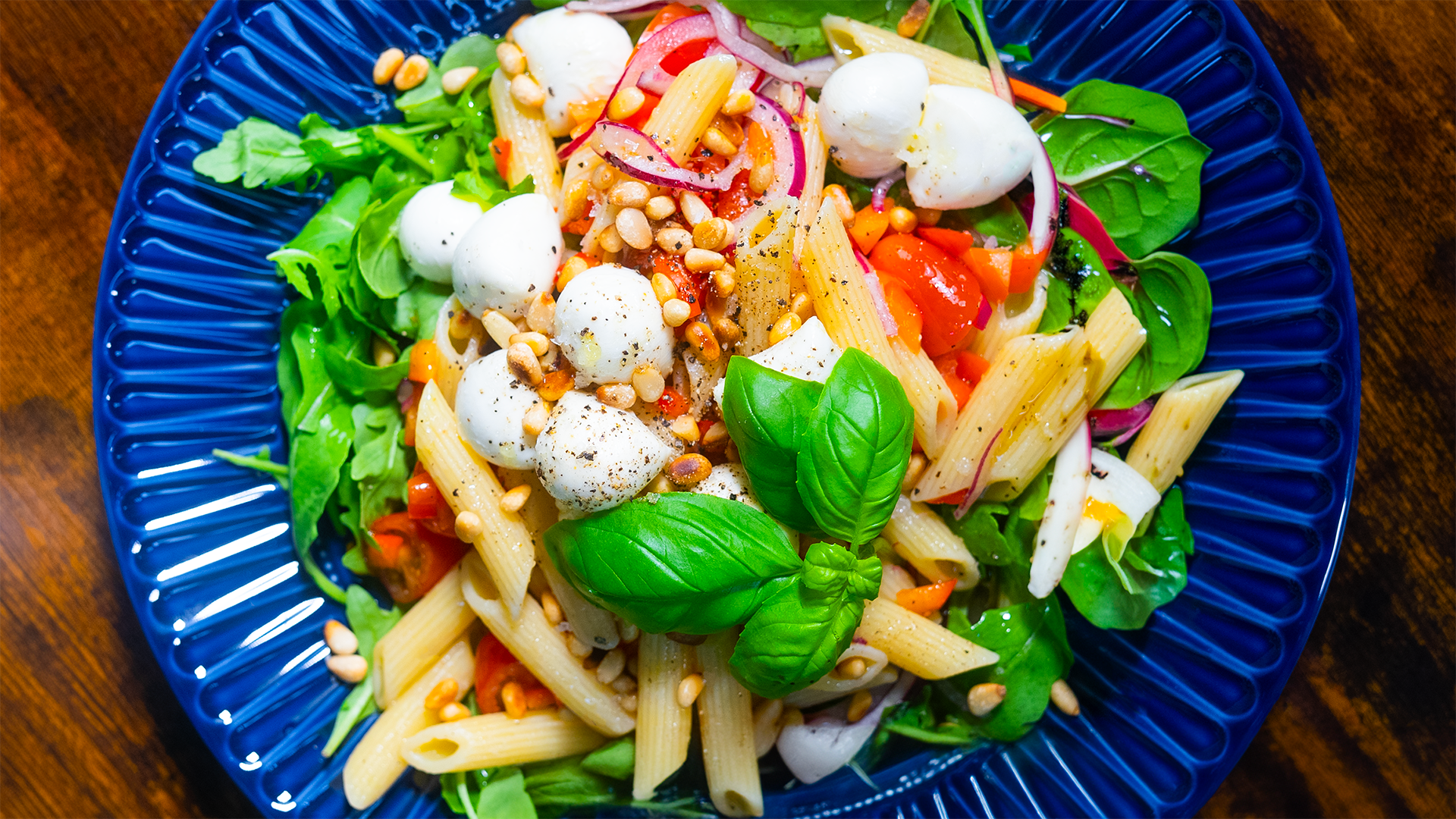 Mediterraner Nudelsalat mit Mozzarella, Rucola, Kirschtomaten und Pinienkernen. Lecker angerichtet auf einem blauen Teller auf dunklem Holztisch