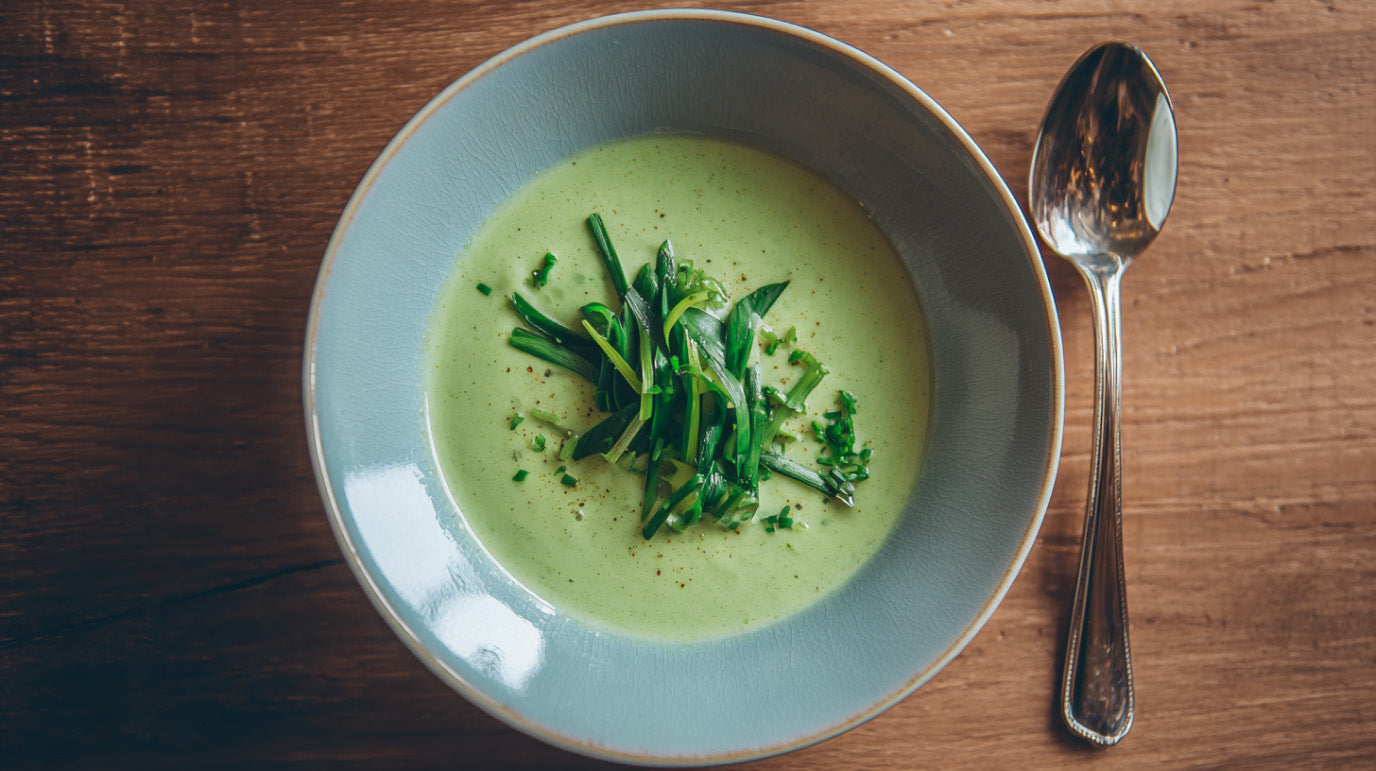 Eine hellgrüne, cremige Bärlauchsuppe in einer blaugrauen Keramikschale, garniert mit frischen Bärlauchstreifen und Pfeffer, serviert auf einem dunklen Holztisch mit einem Silberlöffel daneben.