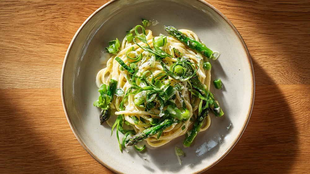 Draufsicht auf eine helle Keramikschale mit Frühlingspasta, bestehend aus Spaghetti in cremiger Sauce, grünem Spargel und feinen Frühlingszwiebelringen auf einem Holztisch.