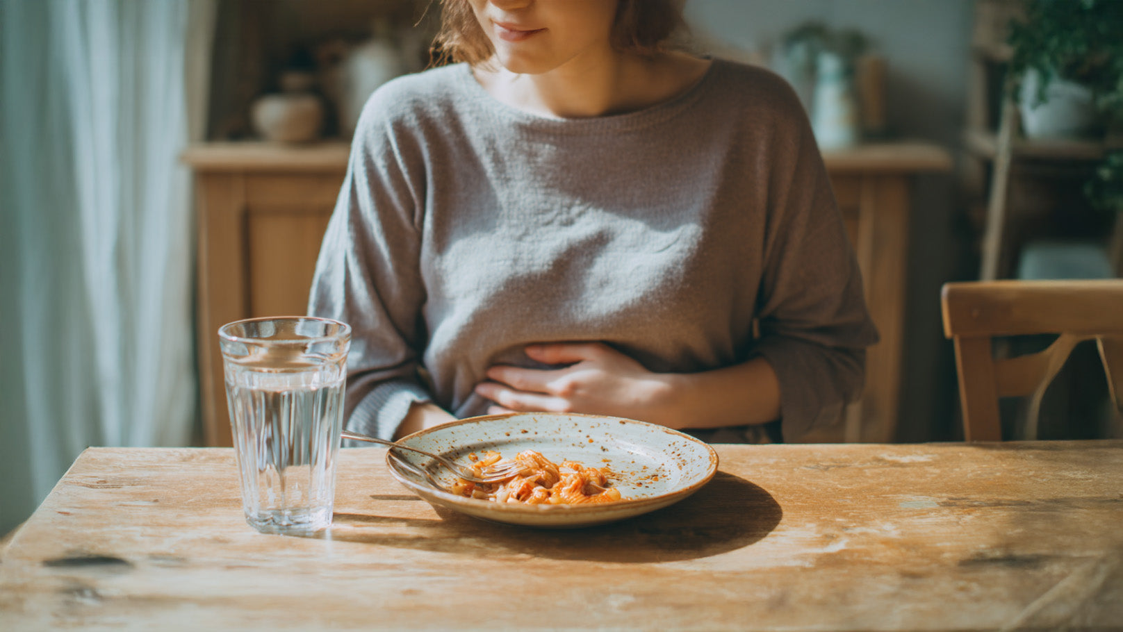 Person sitzt nach dem Essen an einem Tisch, hält sich den leicht gewölbten Bauch, vor ihr ein Teller mit Resten von roter Pastasauce und ein halbvolles Glas Wasser.