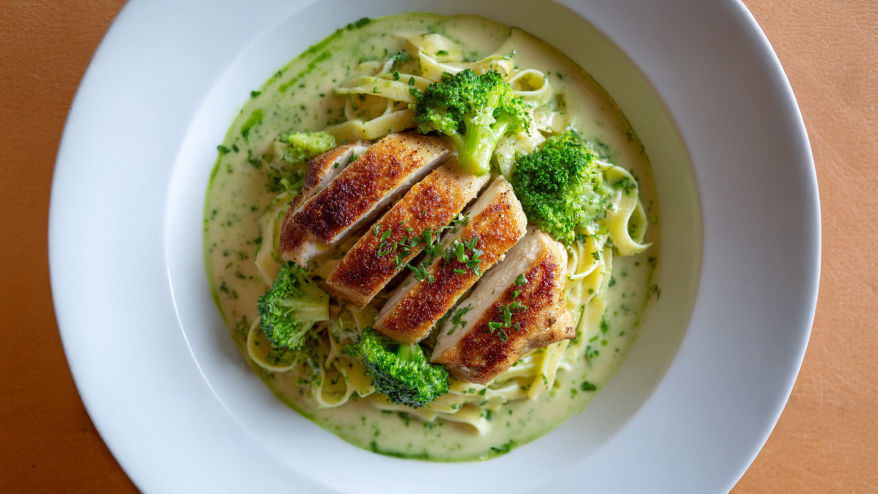 Hähnchenfilet mit Parmesankruste auf cremigen Bandnudeln mit Brokkoli, angerichtet auf weißem Teller mit Kräutersoße