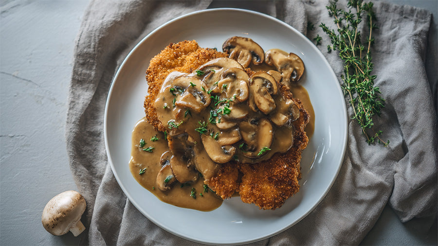 Eine Draufsicht auf einen weißen Teller mit zwei goldbraun panierten Jägerschnitzeln, reichlich bedeckt mit einer cremigen Pilz-Rahm-Sauce und frischem Thymian. Der Teller steht auf einem hellgrauen Leinentuch, daneben liegt ein frischer Champignon und ei