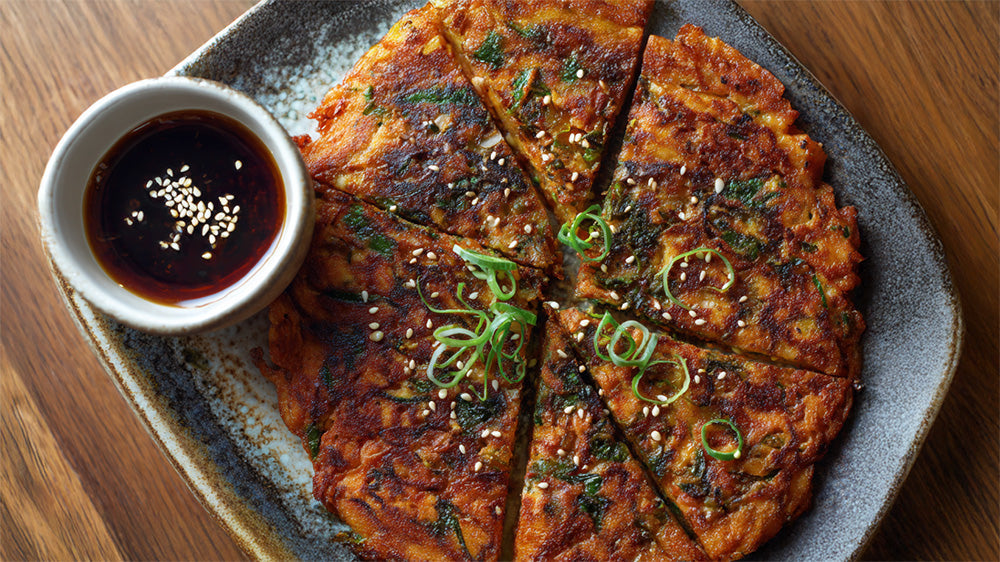 Draufsicht auf einen in Stücke geschnittenen, knusprigen Kimchi-Pfannkuchen (Kimchijeon), garniert mit Lauchzwiebeln und Sesam, serviert mit Dip auf einem dunklen Teller.