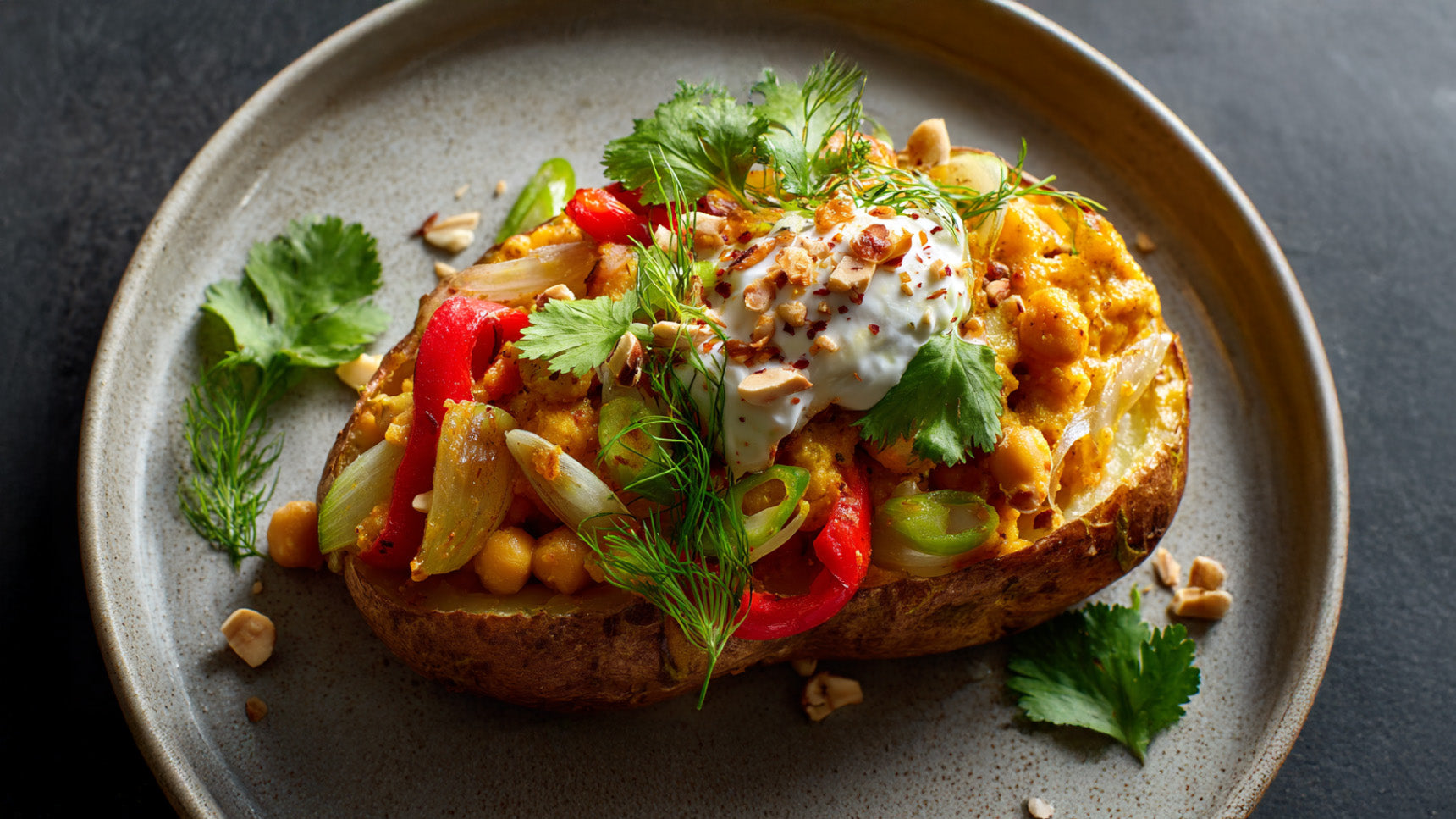 Eine halbierte Ofenkartoffel auf einem grauen Keramikteller, reichlich gefüllt mit Kichererbsen, roten Paprikastreifen und Frühlingszwiebeln. Getoppt mit einem Klecks Sauerrahm, gehackten Nüssen und frischem Koriander vor dunklem Hintergrund.