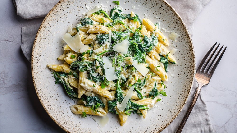 Draufsicht auf einen Teller Penne-Pasta in einer cremigen Spinat-Ricotta-Sauce, garniert mit Parmesan-Hobeln und frischen Kräutern auf einem hellen Untergrund.