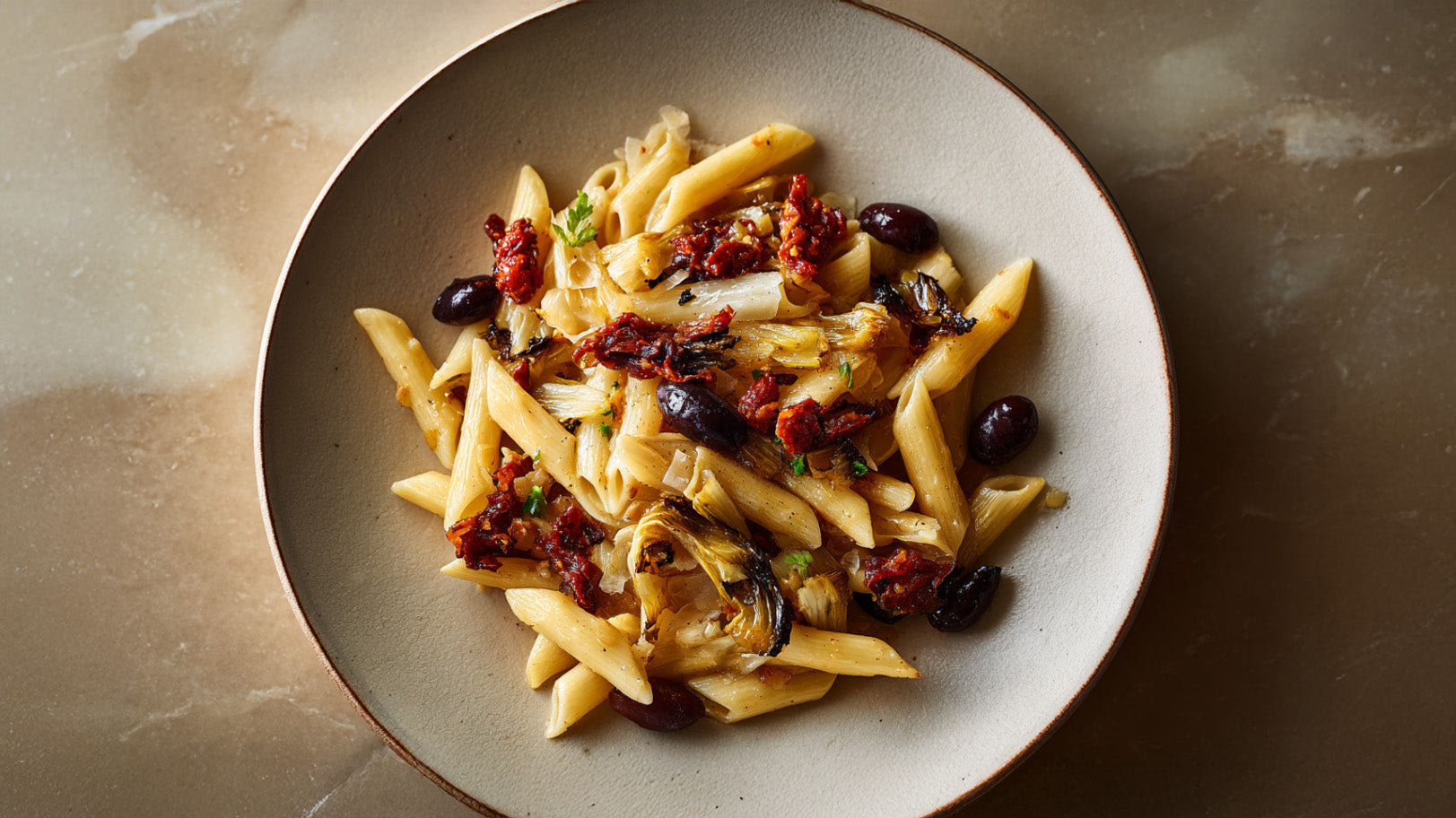 Penne mit gebratenem Chicorée mit getrockneten Tomaten und schwarzen Oliven auf einem hellen Teller, angerichtet als winterliches Pastagericht.