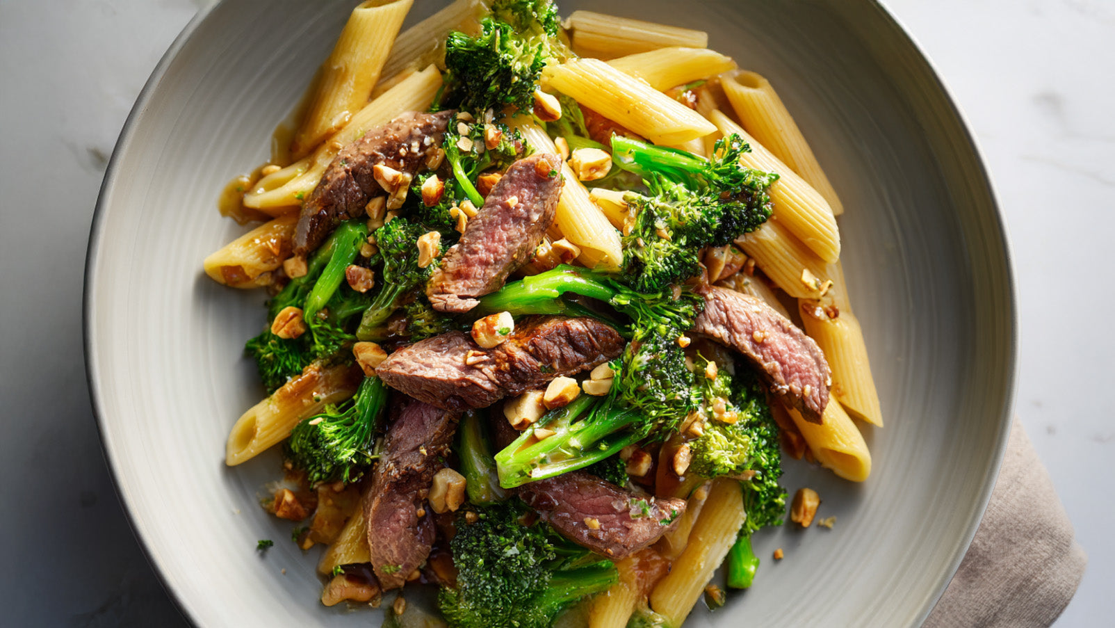 Eine graue Servierschale mit einer Steak-Brokkoli-Pfanne, bestehend aus Penne-Pasta, zarten Rindersteak-Streifen, gebratenem Brokkoli und gehackten Haselnüssen.