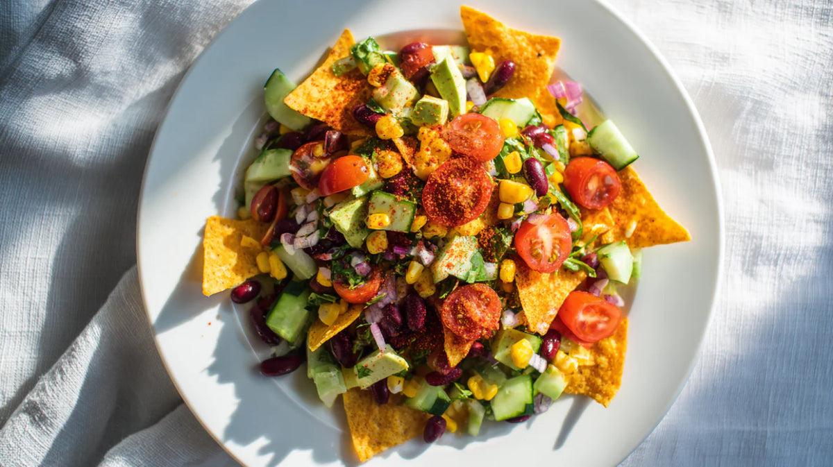 Frischer Tortilla-Salat mit Avocado, Tomaten, Gurken, Mais und Kidneybohnen auf einem weißen Teller, serviert auf einem hellen Tuch, knackige Tortilla-Chips und bunte Zutaten, sommerlicher Tortilla-Salat als leckerer Snack.