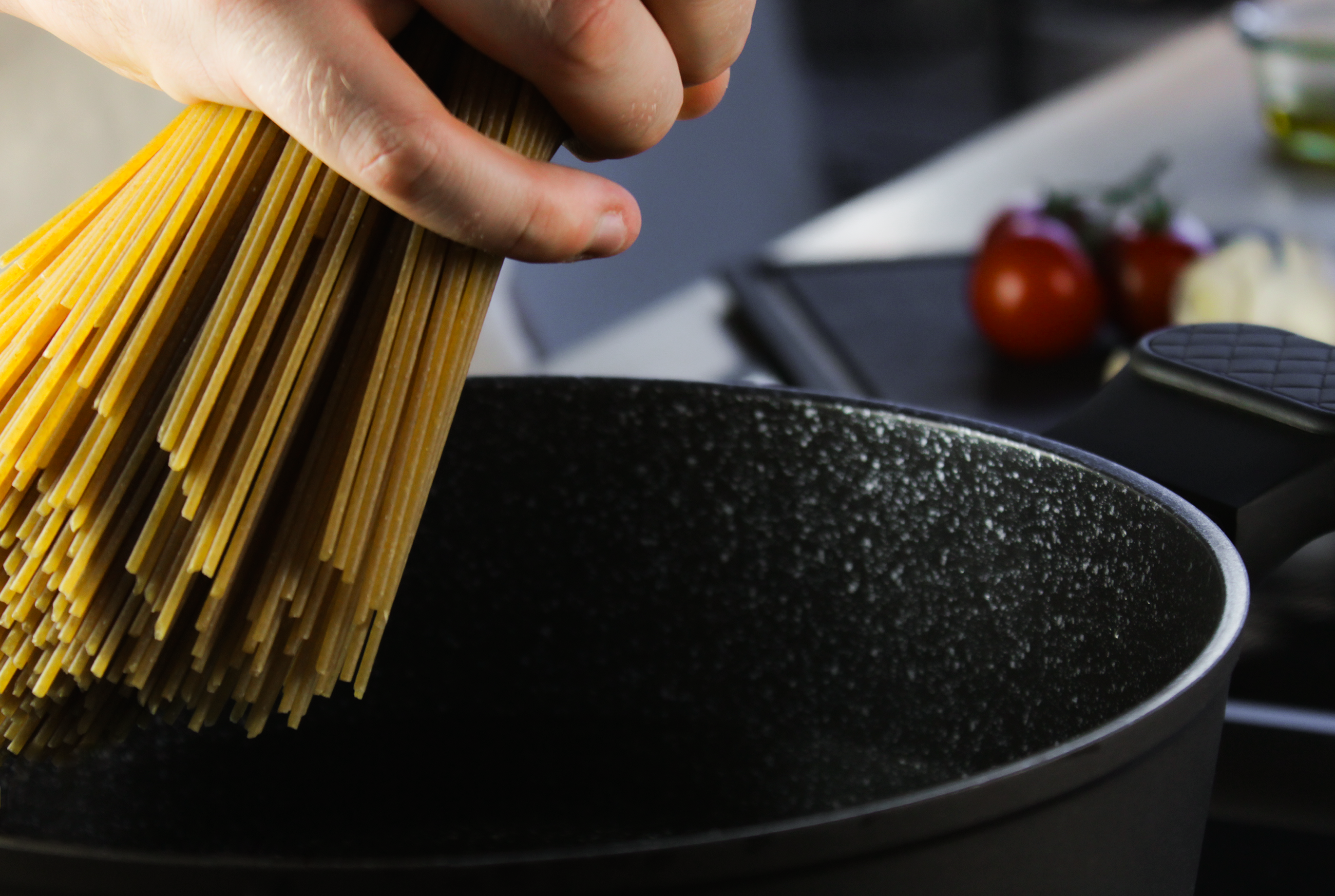 Eine Hand, die Spaghettis in einen FIVI Topf legen wollen mit Tomaten im Hintergrund liegend