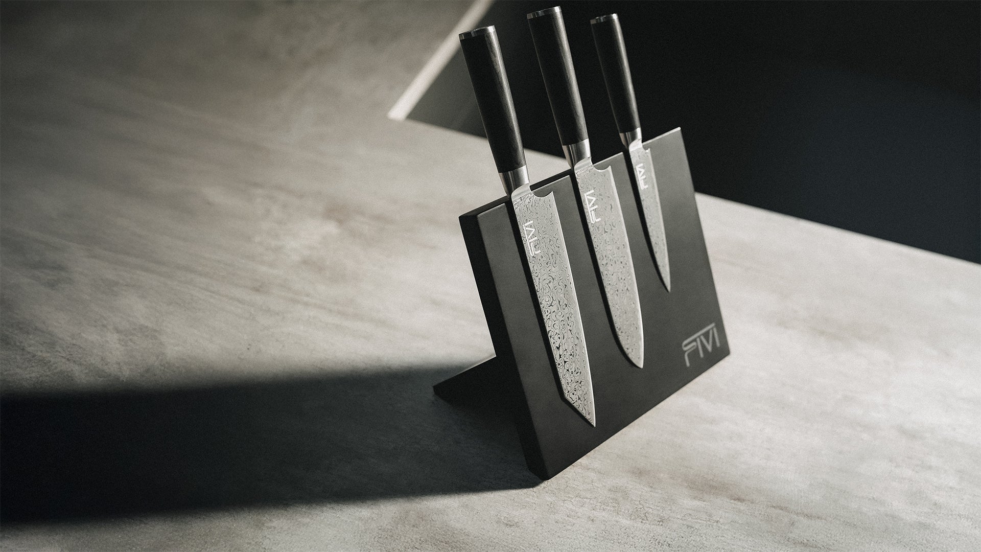 Drei hochwertige FIVI Damastmesser mit dunklen Holzgriffen auf dem schwarzen magnetischen Fivi Messerblock aus Holz vor einem minimalistischen Hintergrund.
