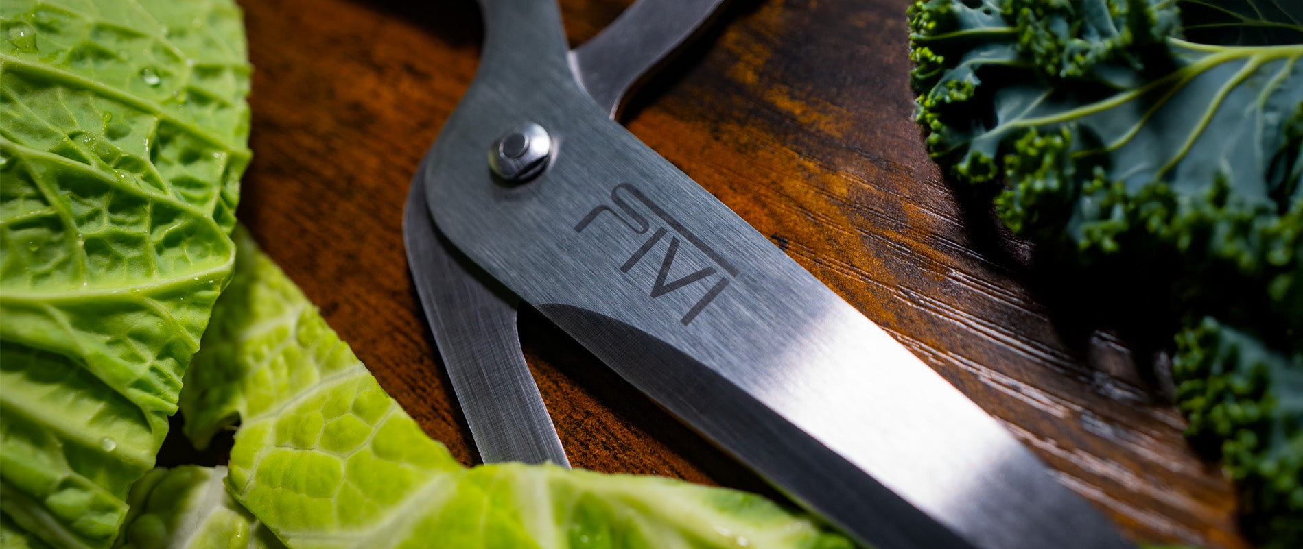 Das FIVI Logo auf der Pizzaschere "Pepe" ist im Fokus und liegt auf einem braunen Untergrund. An den Seiten, rechts und links, liegen Salatblätter.