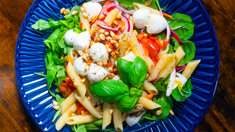 Mediterraner Nudelsalat mit Mozzarella, Rucola, Kirschtomaten und Pinienkernen. Lecker angerichtet auf einem blauen Teller auf dunklem Holztisch