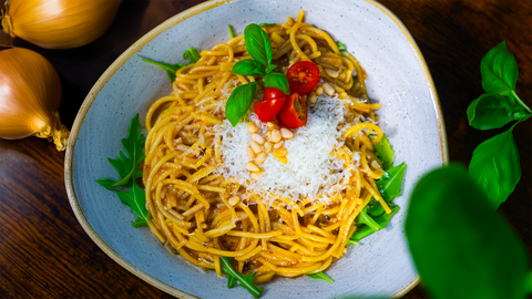 Eine Schüssel mit karamellisierter Zwiebelpasta, garniert mit Kirschtomaten, Basilikum, Pinienkernen und Parmesan, zubereitet in der Schmorpfanne ALMAR, auf einem Tisch mit Zwiebeln und frischem Basilikum im Hintergrund.