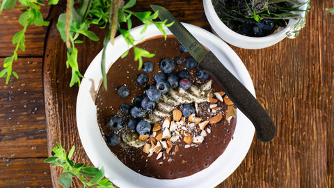 Schoko Bananen Bowl mit Chiasamen, Heidelbeeren und gehackten Mandeln, angerichtet in einer weißen Schale, auf einem Holztisch mit dem Schneidemesser ANIKA von FIVI.