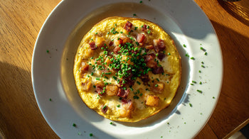 Bauernomelette mit goldgelber Eiermasse, knusprigen Kartoffelwürfeln, Speck und frischem Schnittlauch auf weißem Teller, angerichtet auf Holztisch