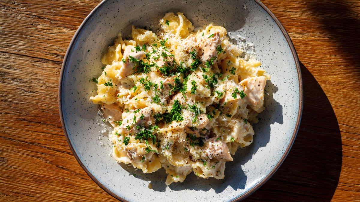 Draufsicht auf eine Portion Cheesy Chicken Pasta in einer grauen Keramikschüssel. Die Pasta ist mit einer cremigen Käsesauce, gebratenen Hähnchenstücken, viel geriebenem Parmesan und frischer Petersilie angerichtet. Das Gericht steht auf einem Holztisch i
