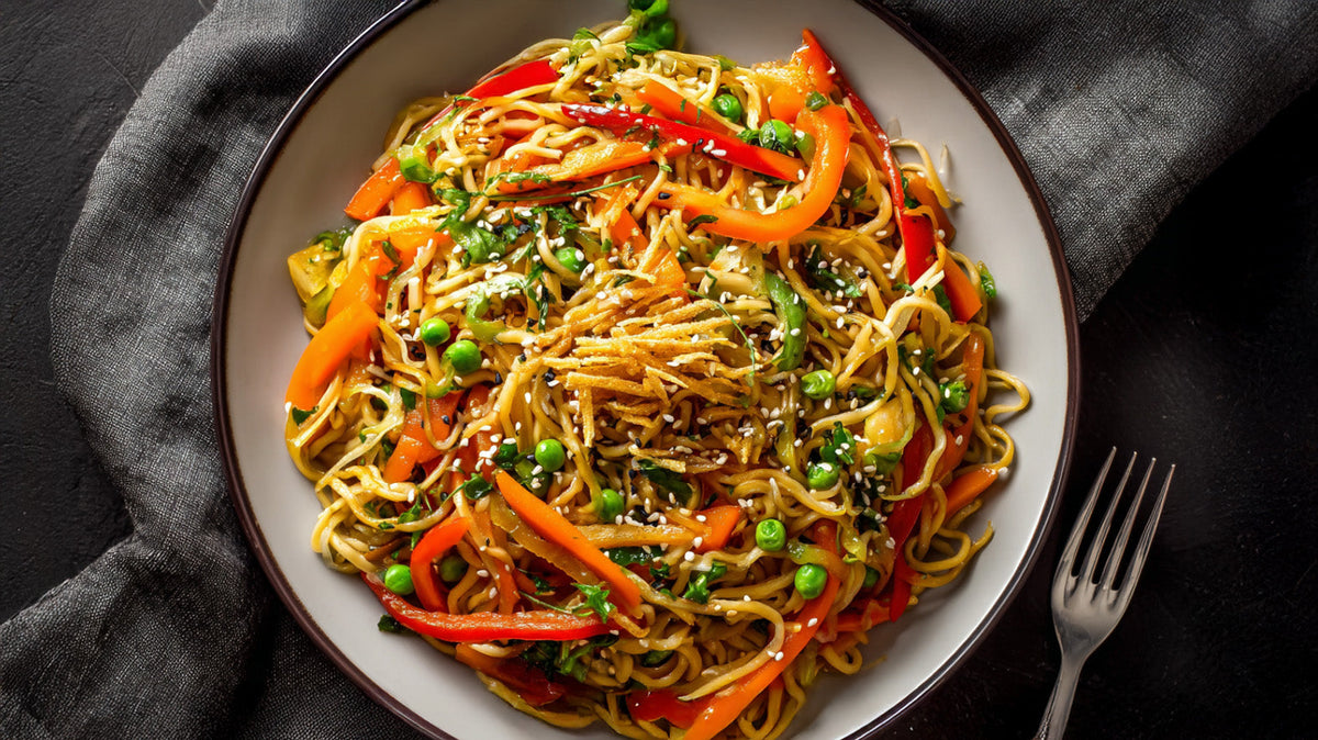 Chinesische Bratnudeln mit Gemüse in einer Schale, angerichtet mit Paprika, Erbsen, Sprossen und Sesam auf dunklem Untergrund