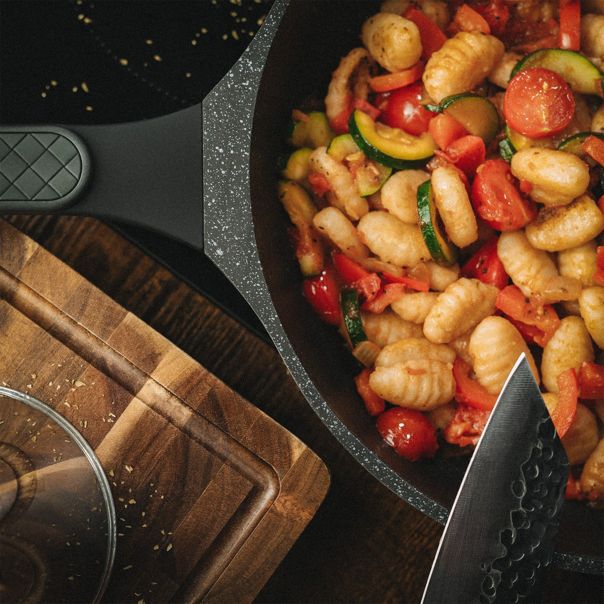 Detailaufnahme von gebratenen Gnocchi mit Kirschtomaten und Zucchini in einer beschichteten Pfanne neben einem Schneidebrett.