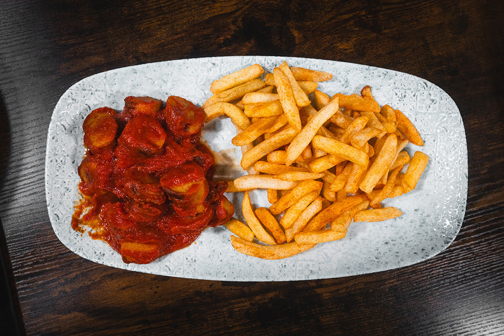 Ein rechteckiger weißer Teller mit Currywurst in roter Currysauce auf der linken Seite und einer Portion goldbrauner Pommes auf der rechten Seite, auf einem dunklen Holztisch.