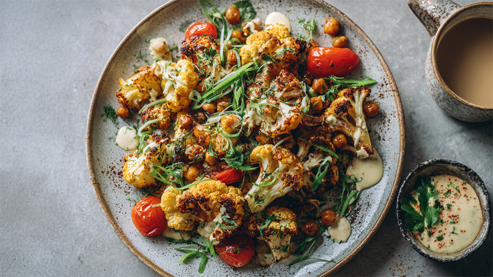 Draufsicht auf einen großen, rustikalen Teller mit gebratenem Blumenkohl, gerösteten Kichererbsen und Kirschtomaten, garniert mit Kräutern und Sauce. Daneben stehen zwei kleine Schälchen.