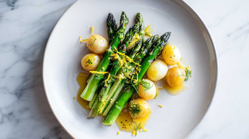 Draufsicht auf grünen Spargel mit kleinen Drillingen, garniert mit Zitronenabrieb, frischem Dill und Zitronenbutter auf einem weißen Teller.