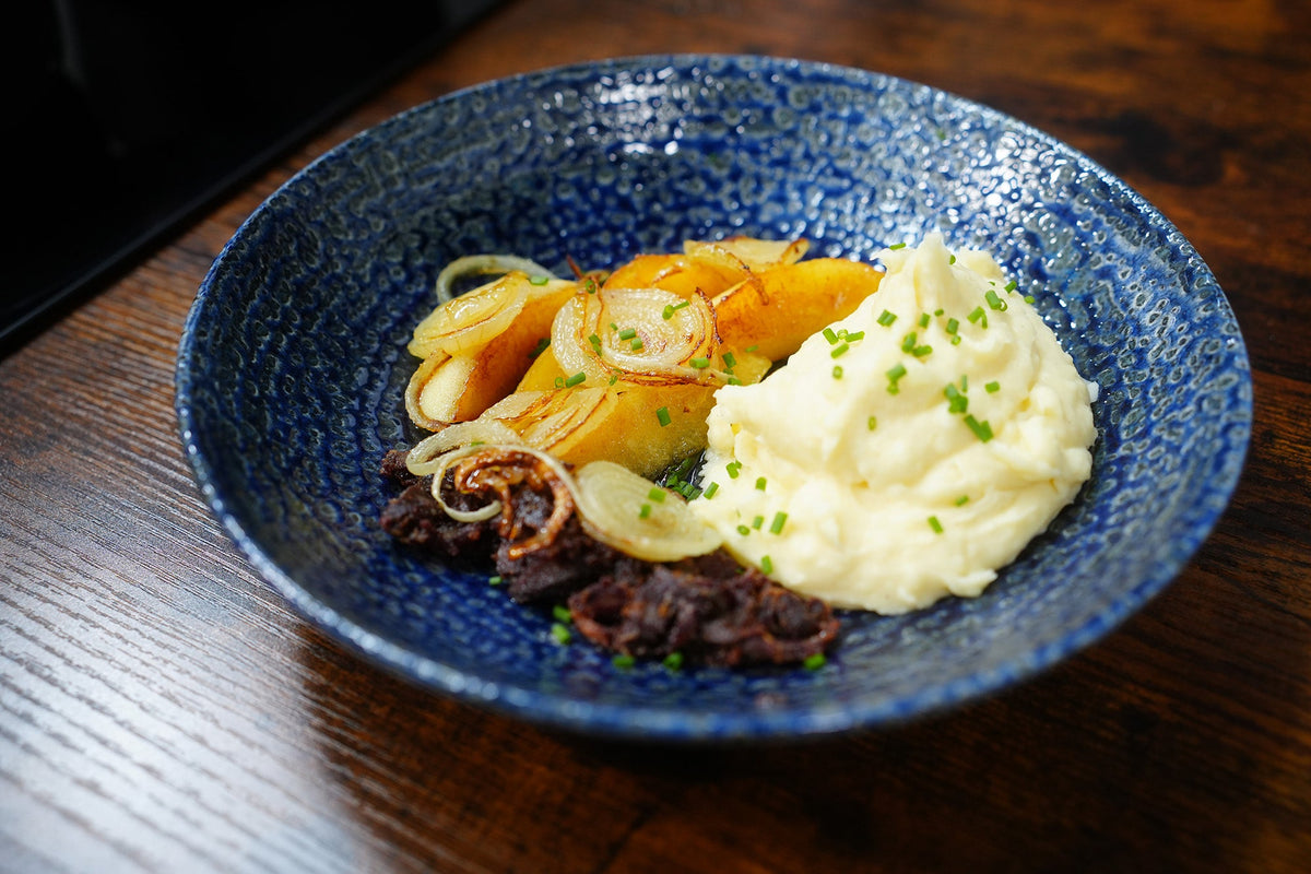 Traditionelles Himmel & Äd angerichtet in einer blauen Keramikschale: cremiges Kartoffelpüree mit Schnittlauch, gebratene Apfelspalten, knusprige Zwiebelringe und herzhafte Blutwurst.