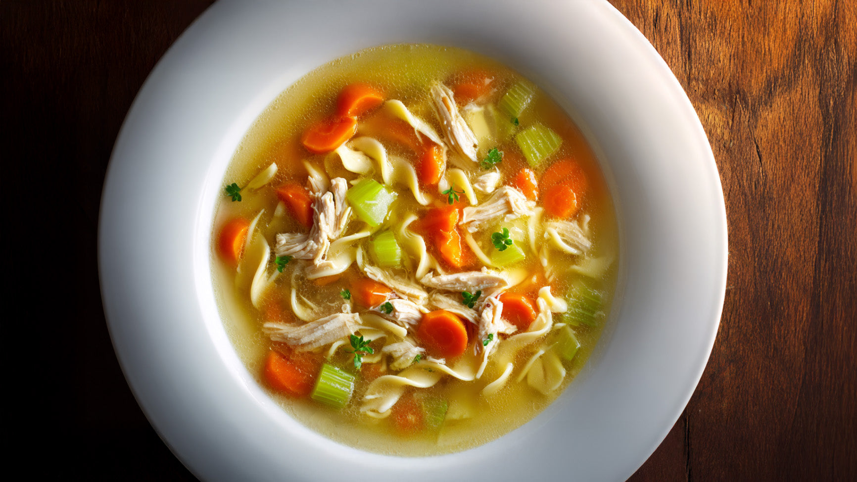Hühnersuppe mit Nudeln in weißer Schale, klare Brühe mit zartem Hähnchenfleisch, Karotten, Sellerie und frischer Petersilie – von oben fotografiert