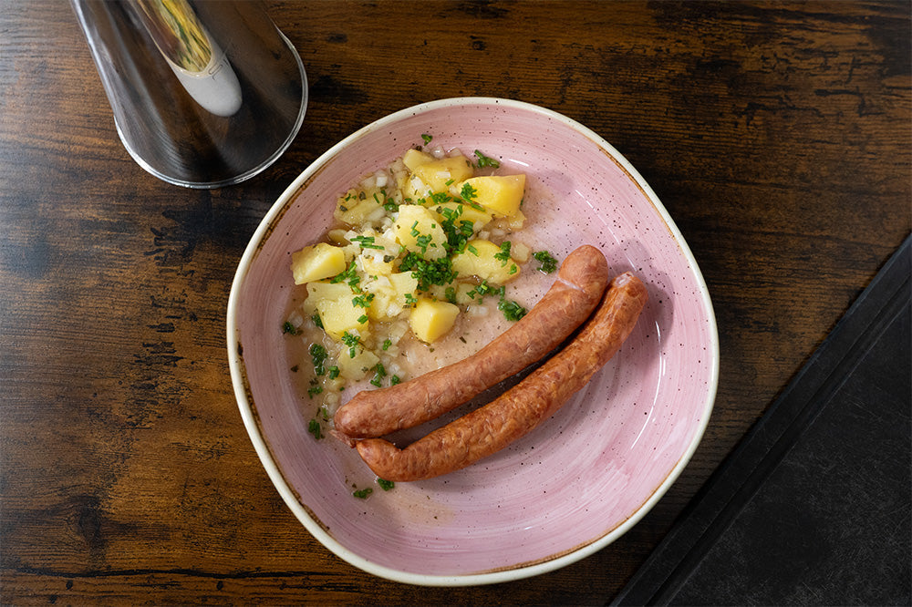 Nahaufnahme eines rosa Tellers mit schwäbischem Kartoffelsalat (mit Zwiebeln und Schnittlauch) und zwei Saitenwürstchen (Wiener Würstchen) auf einem braunen Holztisch.