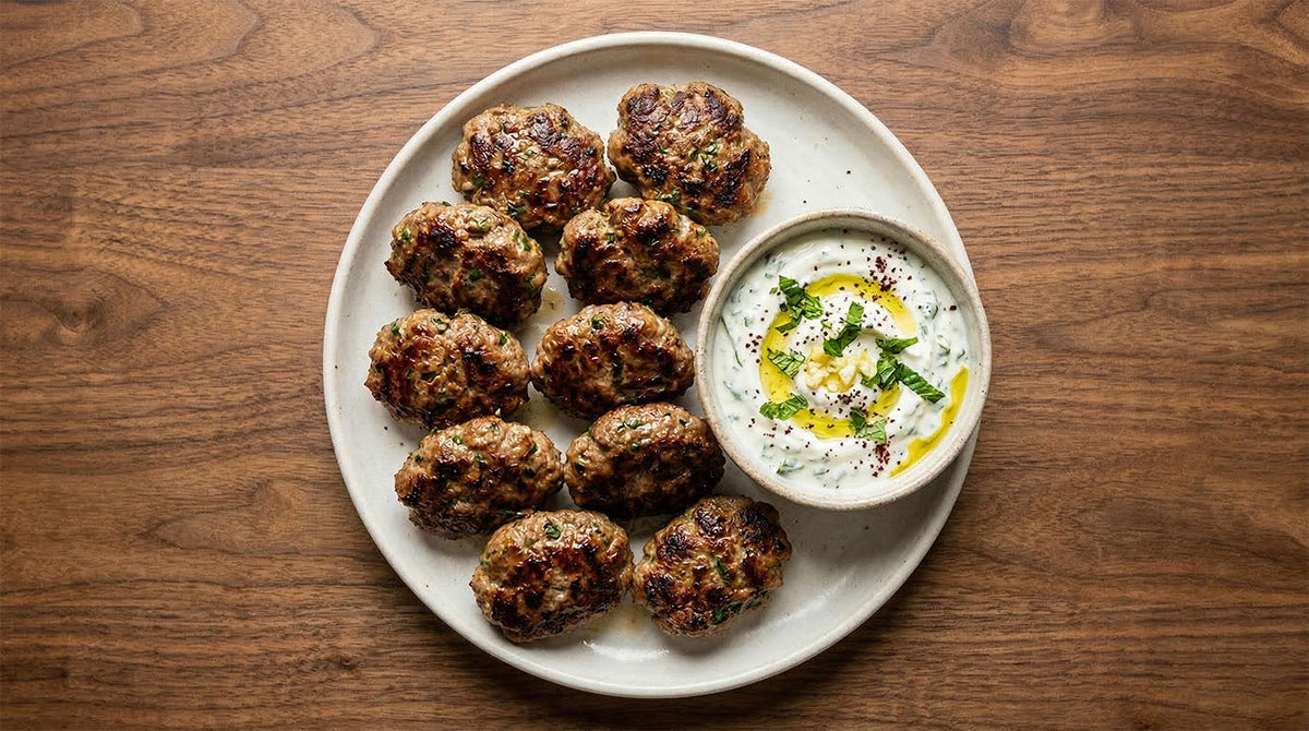 Draufsicht auf einen hellen Keramikteller mit saftig gebratenen Köfte und einer Schale Joghurt-Dip mit Olivenöl und Kräutern auf einem dunklen Holztisch.