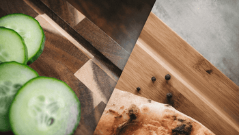 Frische Gurkenscheiben auf einem dunklen Akazien-Schneidebrett links und ein Stück gebackenes Fladenbrot mit schwarzen Pfefferkörnern auf hellem Bambus-Schneidebrett rechts, Zutaten für ein rustikales Gericht.