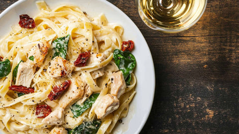 Cremige Pasta mit Hähnchen, getrockneten Tomaten und Spinat auf einem weißen Teller, serviert mit frisch geriebenem Parmesan und einem Glas Weißwein.