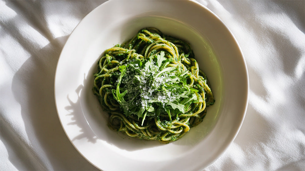 Draufsicht auf einen tiefen Teller mit Pasta Verde, garniert mit frischem Rucola und geriebenem Parmesan auf einer weißen Tischdecke.