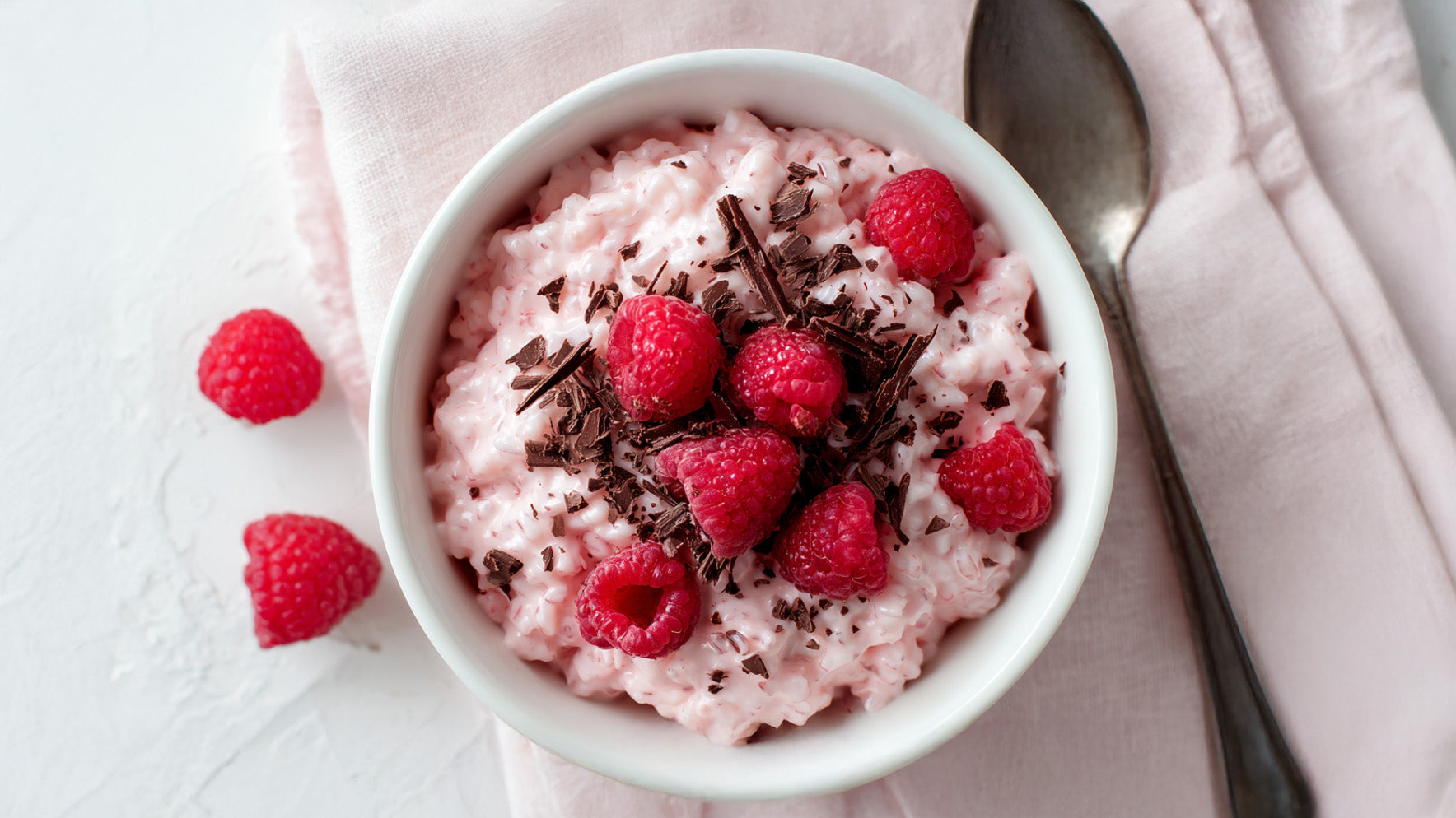 Pink Milchreis mit Himbeeren und Schokolade, cremig angerichtet in weißer Schale, getoppt mit frischen Himbeeren und feinen Zartbitterschokoraspeln.