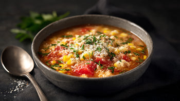 Pizzasuppe in Schale mit Mais, Tomatenstücken, Kräutern und geriebenem Käse, serviert auf dunklem Untergrund mit Löffel daneben.