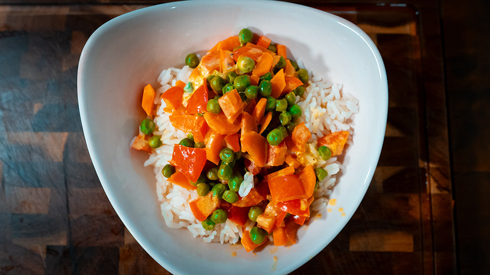 Draufsicht auf eine weiße, geschwungene Keramikschale, gefüllt mit weißem Reis und einer großzügigen Portion Gemüse-Sauce aus Erbsen und gewürfelten Karotten in einer cremigen, hellen Sauce.