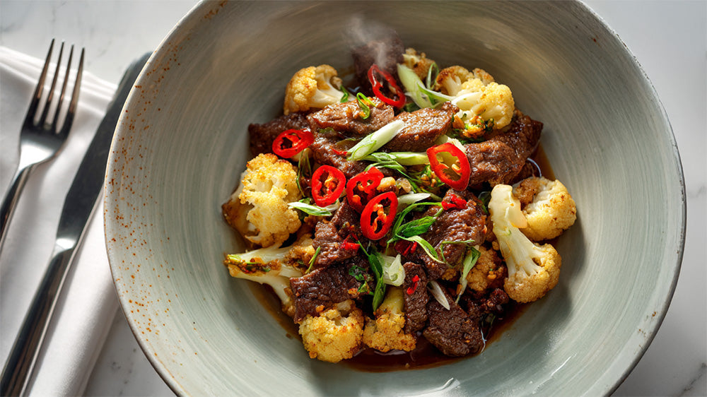 Draufsicht auf gebratenes Rindfleisch mit Blumenkohl, garniert mit Chili und Frühlingszwiebeln, in einer Schüssel neben Besteck.
