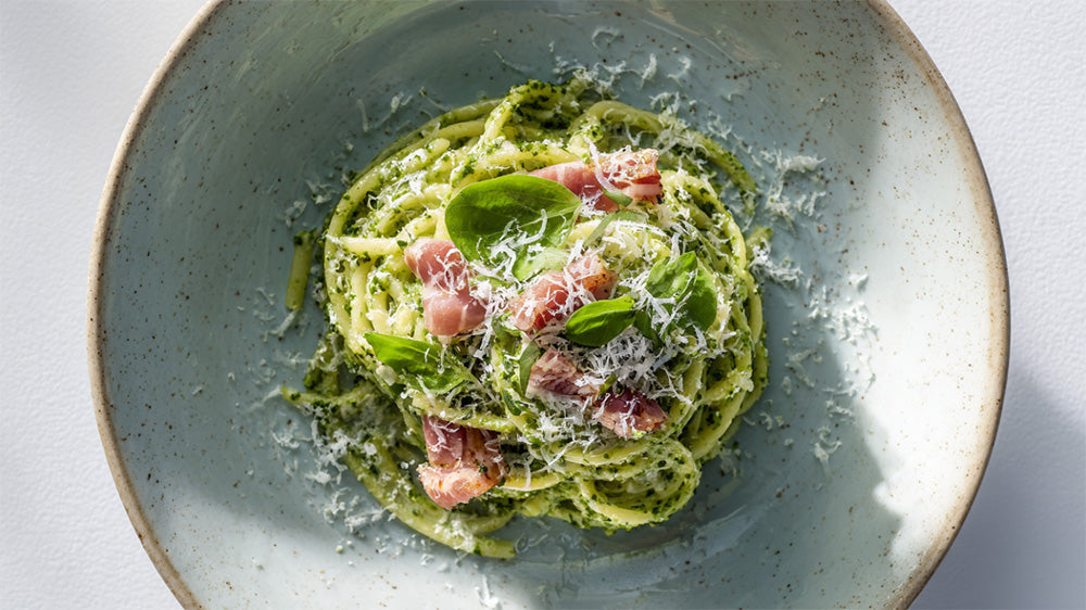 Nahaufnahme von Spaghetti in einer leuchtend grünen Bärlauch-Pesto-Sauce, garniert mit gewürfeltem Schinken, frisch geriebenem Parmesan und Basilikumblättern in einer rustikalen Keramikschüssel.