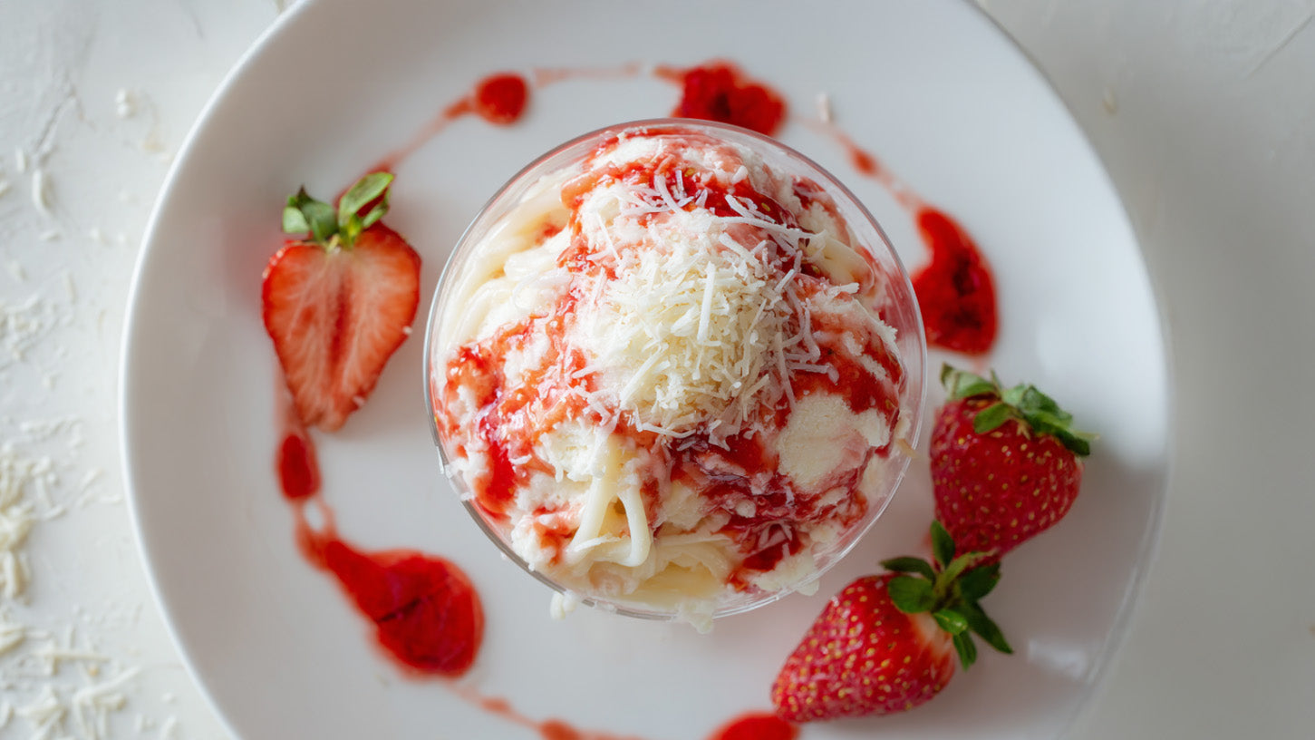 Spaghetti Eis Glasdessert mit Erdbeersauce und weißer Schokolade in Dessertglas angerichtet, umgeben von frischen Erdbeeren auf weißem Teller