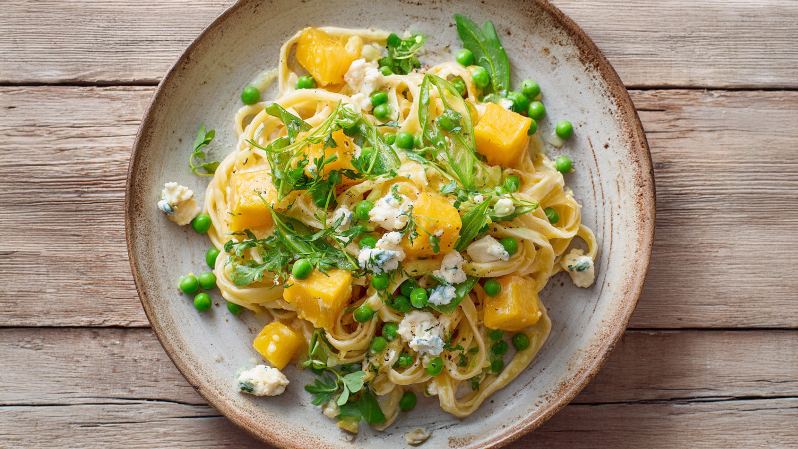 Cremige Pasta mit Steckrübe, Erbsen, Zuckerschoten und Ziegenfrischkäse, von oben fotografiert, serviert auf einem rustikalen Teller mit frischen Kräutern und Kresse, natürliche Beleuchtung auf Holztisch.