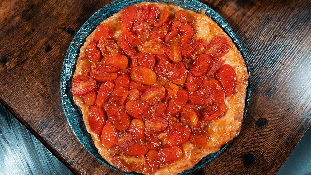 Eine frisch gebackene, runde Tomaten-Tarte-Tatin auf einem blau-gemusterten Teller, von oben fotografiert. Die Oberfläche ist dicht belegt mit glänzenden, karamellisierten roten Kirschtomaten auf einem goldbraunen Teigboden. Der Teller steht auf einem dun