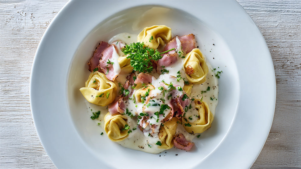 Draufsicht auf Tortellini alla panna mit Schinken und viel Petersilie in einem weißen Teller auf Holztisch.