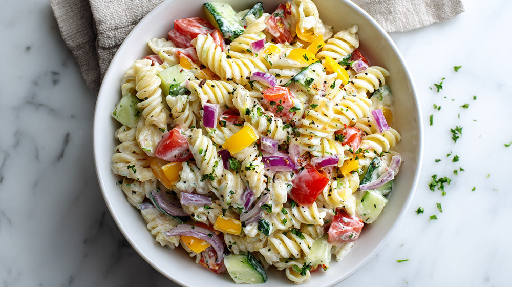 Draufsicht auf eine weiße Schüssel mit Tzatziki-Nudelsalat aus Spirelli, Tomaten, Gurken, gelber Paprika und roten Zwiebeln auf einem hellen Marmorhintergrund.