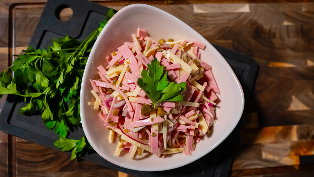 Draufsicht auf eine weiße Schüssel mit Schweizer Wurstsalat aus feinen Lyoner-Streifen, Käse, roten Zwiebeln und Gurken, garniert mit Petersilie auf einem dunklen Schneidebrett.