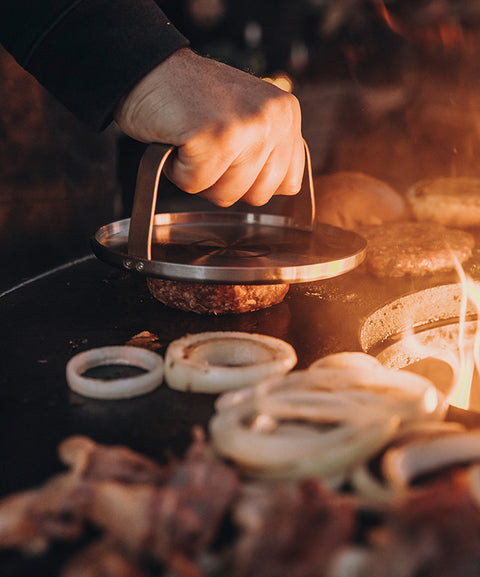 Burgerpresse in Aktion bei der Zubereitung perfekter Burger-Patties auf einem Grill, mit frischen Zutaten im Hintergrund – FIVI Moesta.