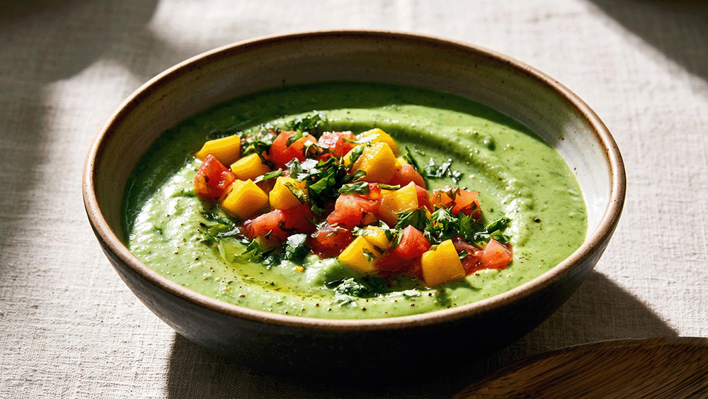 Cremige Sommer-Suppe mit Erbsen und Koriander in rustikaler Schale, angerichtet mit frischem Mango-Tomaten-Topping im warmen Sonnenlicht.