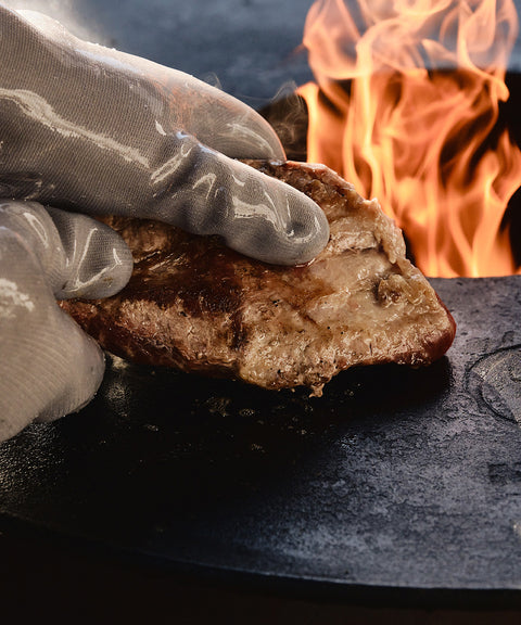 Anwendung der hitzefesten Handschuhe von FIVI Moesta beim sicheren Umgang mit heißen Grillutensilien, Grill im Hintergrund.