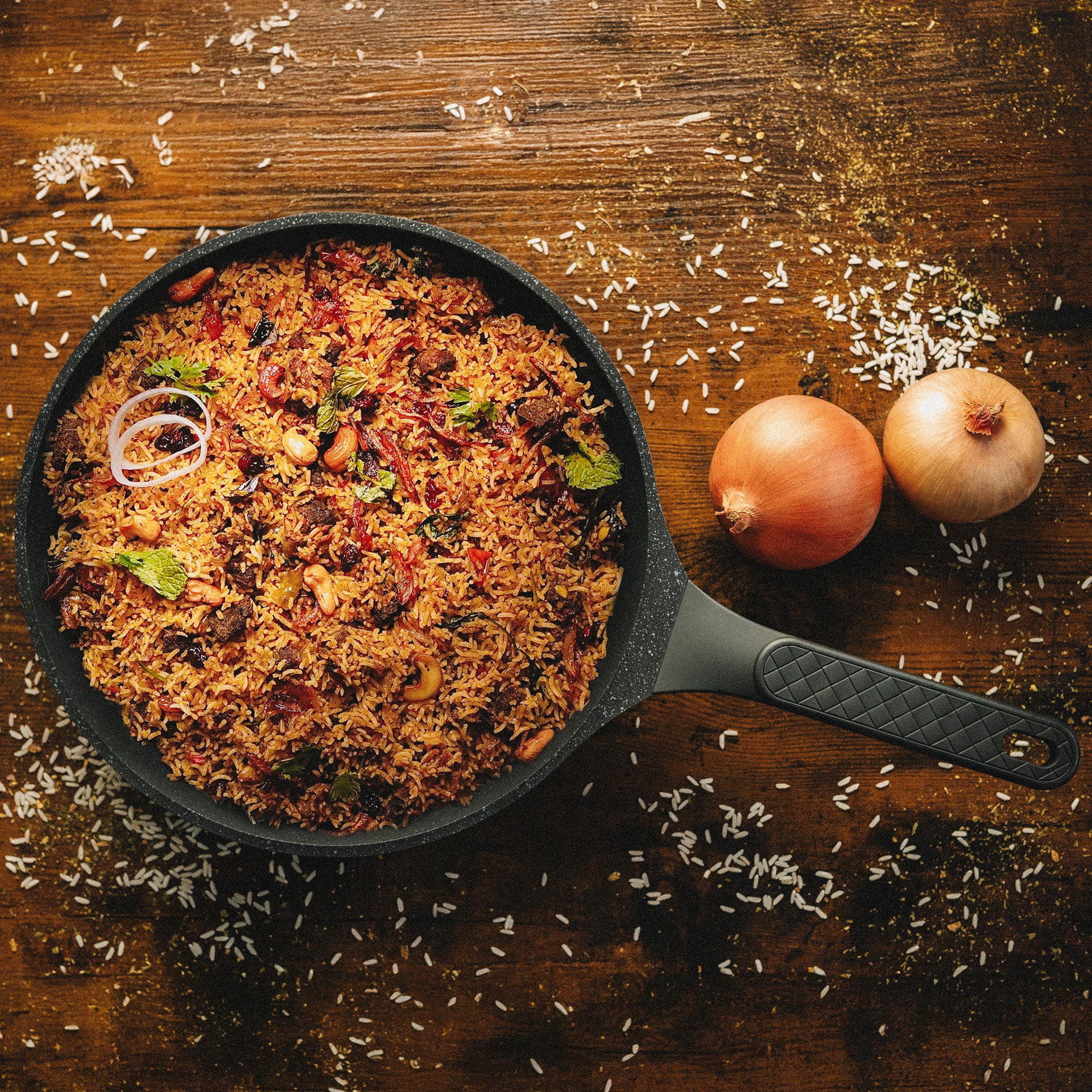 Hochwertige beschichtete Bratpfanne Vanessa mit einem aromatischen Reisgericht neben ganzen Zwiebeln und verstreuten Reiskörnern.