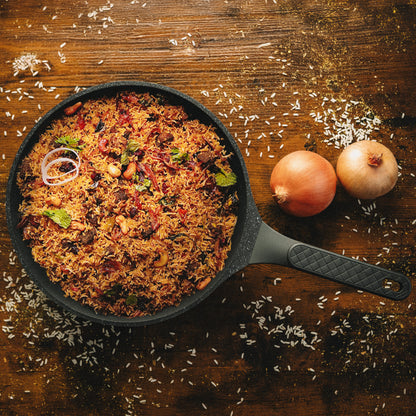 Hochwertige beschichtete Bratpfanne Vanessa mit einem aromatischen Reisgericht neben ganzen Zwiebeln und verstreuten Reiskörnern.
