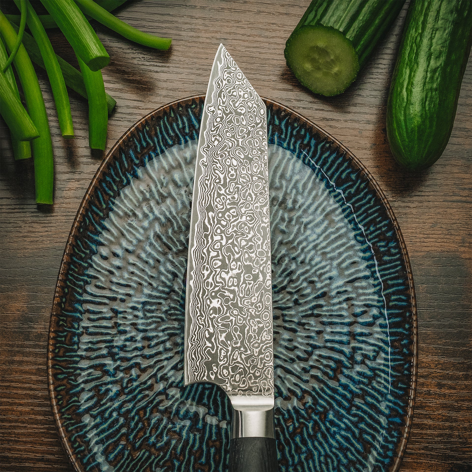 Ästhetische Draufsicht des Yuno Damastmessers auf einem blauen Teller neben frischen Gurken.