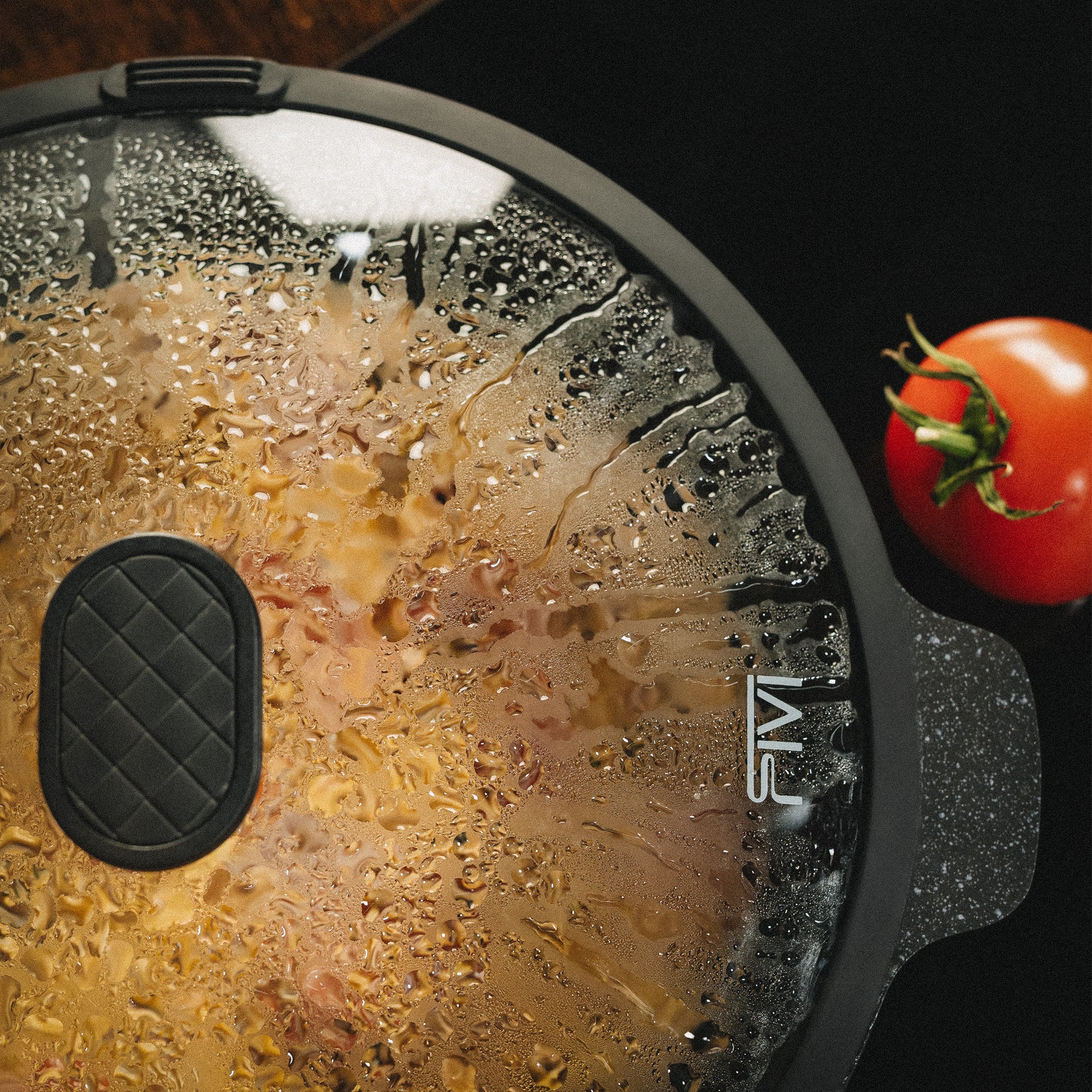 Glasdeckel der Schmorpfanne Almar mit Dampfauslass und Kondenswasser über einer brutzelnden Pfanne, daneben eine frische Tomate auf dunkler Arbeitsplatte.